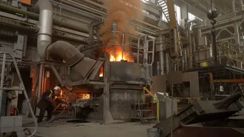 BBC News / Kevin Church Flames emerging from the top of the arc furnace as material is added to the molten steel